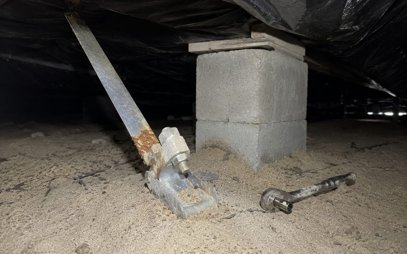 Rusted tie-down strap running to a weathered ground anchor head beside a concrete block pier under a home