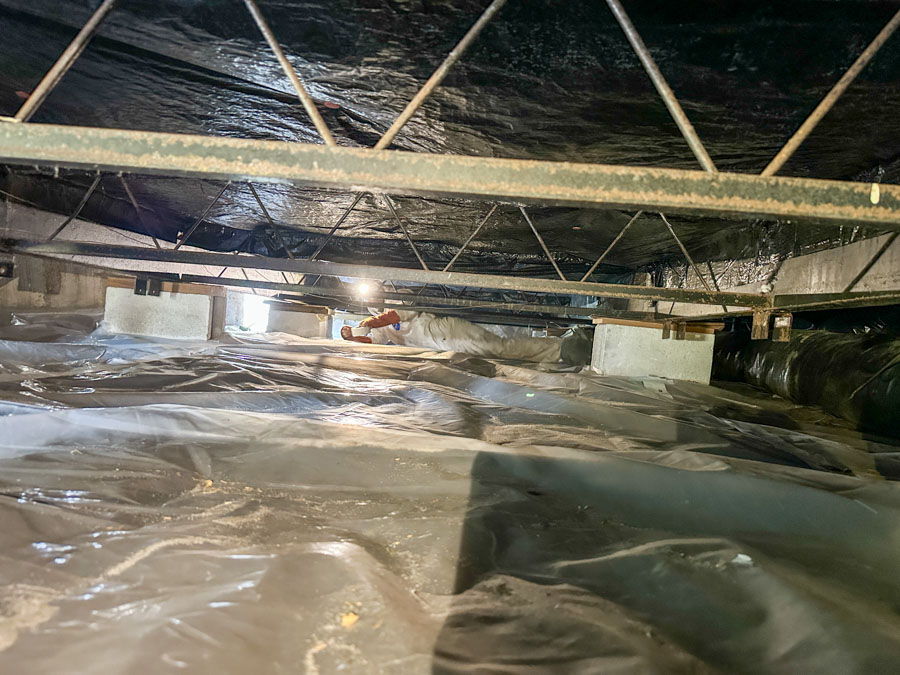 An East Coast Vapor Barrier technician in protective gear works inside a fully encapsulated crawl space, illuminated by a work light, ensuring every seam and corner of the vapor barrier is properly sealed.