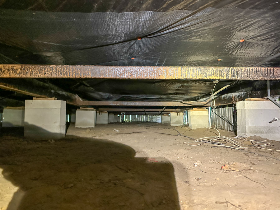 Wide-angle shot of an unencapsulated crawl space featuring exposed steel I-beams, concrete piers, and a dirt floor — illustrating the pre-installation condition before East Coast Vapor Barrier’s moisture control system is applied.