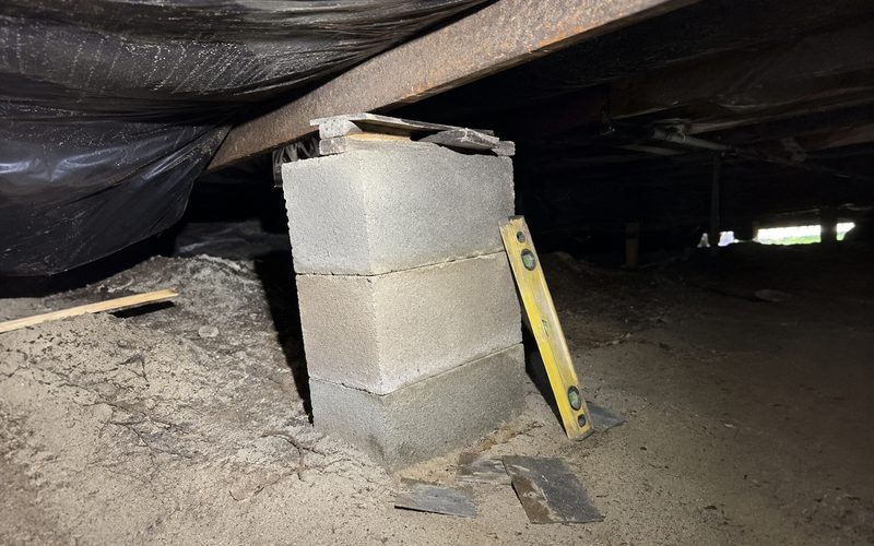 Concrete block pier settled out of level under a home, with the old shims split and falling out of the gap beneath the steel beam
